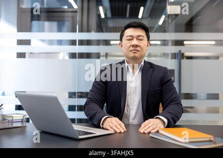 Asian businessman in a formal suit sitting at his modern office desk with closed eyes, practicing mindfulness and stress relief through meditation and calm relaxation during a workday Stock Photo