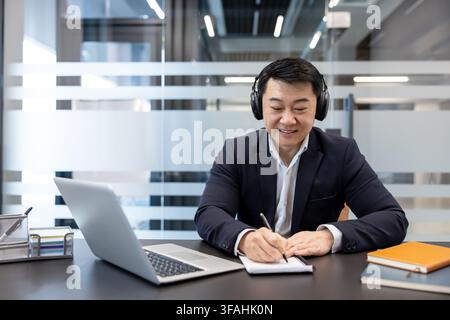 Asian businessman in a suit wearing headphones, smiling while writing notes in a notebook at his desk, working on a laptop in a contemporary office setting Stock Photo