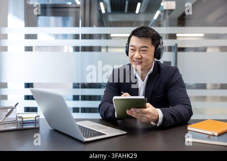 Asian businessman in formal wear sitting at a modern office desk, wearing headphones, watching a webinar on his laptop and attentively taking notes in a notebook Stock Photo