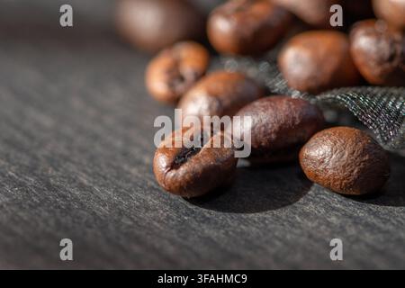 Roasted coffee beans lie scattered on a dark textured surface with shallow focus. Stock Photo