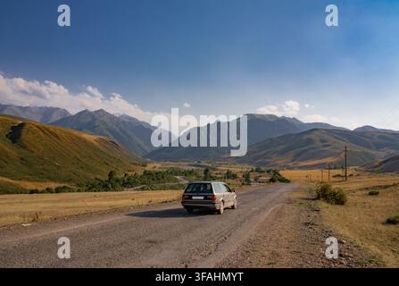 A silver station wagon drives along a rough gravel road through a wide mountain valley, with a river winding through green fields and layered peaks ri Stock Photo
