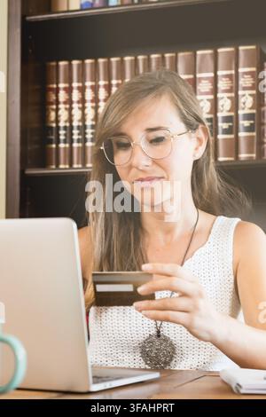 Young woman with glasses holds a bank card while using a laptop at home, illustrating online shopping, digital payment, ecommerce and secure banking. Stock Photo