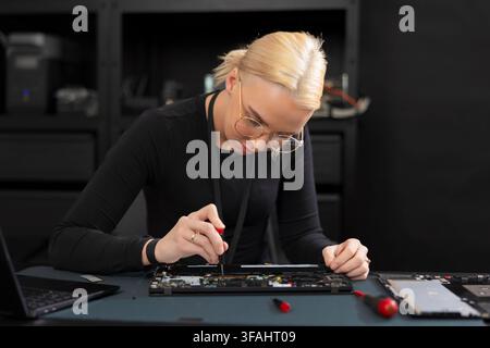 A Caucasian woman in her early 30s, wearing glasses, focuses on hardware repair inside a tech workspace. She uses a precision screwdriver to access th Stock Photo