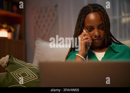 Black Woman Talking on Smartphone and Working on Laptop at Home Stock Photo