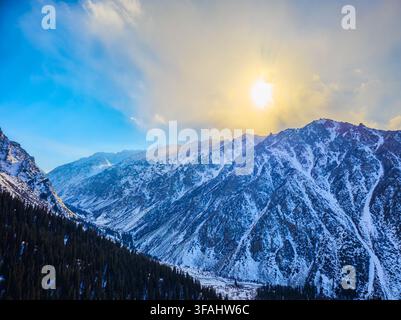Wide view of majestic snow-covered mountains illuminated by sunlight breaking through clouds. Dense coniferous forest blankets the lower slopes, enhan Stock Photo