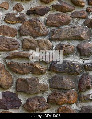 Vertical close-up view of a weathered rock wall surface with large porous stones in variegated brown tones and thick, porous mortar Stock Photo