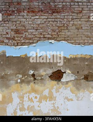 Vertical view of a rundown multi-layer wall surface with peeling paint in several colors revealing cracked concrete with red brick with worn mortar Stock Photo