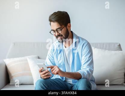 Useful App. Handsome Arab Man In Eyeglasses Using Smatphone At Home Stock Photo