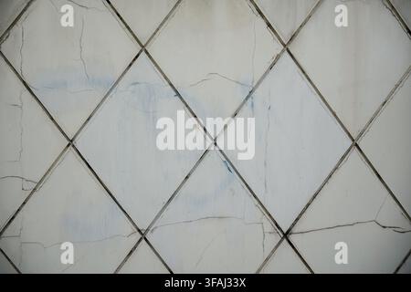 Close up of an old white tiled wall with diagonal square patterns. The ceramic tiles show multiple cracks, damage, and weathered grout lines. Stock Photo