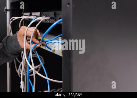 Medium shot view of a technician plugging a blue ethernet cable into a network switch inside a server rack. Stock Photo