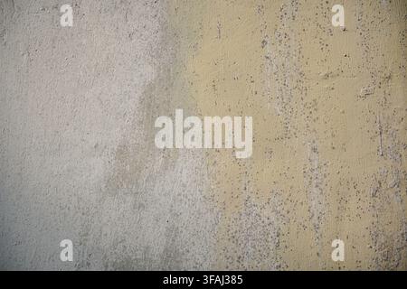 Close up of a textured wall background showing a transition between plain grey concrete and faded, peeling pale yellow paint with dark mold spots. Stock Photo