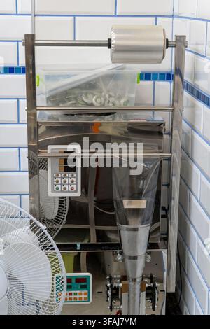 Industrial stainless steel packaging machine with film roll stands in kitchen with white tiled walls, plastic container with green vegetables, and ele Stock Photo