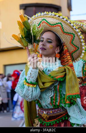 Young woman wears a colorful traditional Mexican costume with a large, decorated hat. She holds corn cobs and greenery, symbolizing harvest or cultura Stock Photo