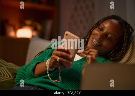Smiling Black Woman Using Smartphone on Sofa at Night Stock Photo