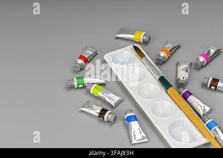 Arrangement of colorful watercolor tubes, a white plastic mixing palette, and a classic artist brush on a minimal gray studio background. Stock Photo