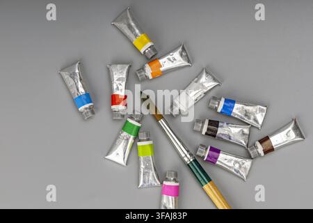 A collection of colorful watercolor paint tubes and a pointed artist paintbrush arranged on a smooth neutral gray background in flat lay perspective. Stock Photo