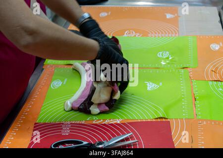 Hands in black protective gloves twist purple white and pink caramel pieces together on silicone workspace sheet with metal scissors visible in foregr Stock Photo