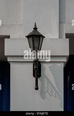 Close-up shot of a vintage black metal street lamp post mounted on a white concrete pillar, casting a defined shadow against the wall. Stock Photo