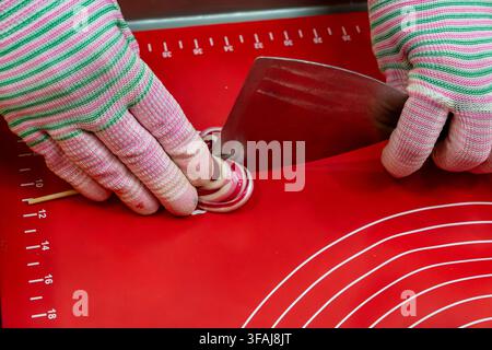 Hands in striped textile gloves hold sharp steel spatula device near round striped caramel candy mass on red silicone measurement mat during artisanal Stock Photo