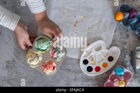 Creative Easter flat lay showing hands arranging colorful marbled Easter eggs in shaving foam inside glass bowls with watercolor paints and craft supp Stock Photo