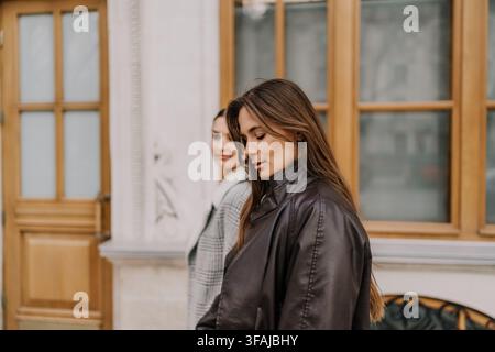 Women coats city walking together in stylish autumn apparel past a classic urban building with large windows during the day Stock Photo