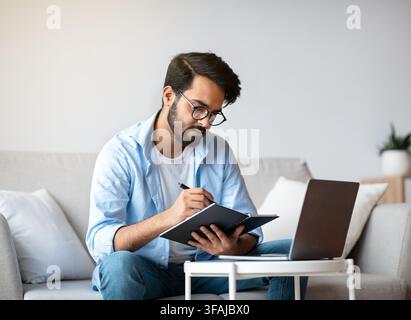 Young Arab Freelancer Man Taking Notes And Working On Laptop At Home Stock Photo