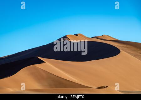 Dramatic view of towering sand dunes under a clear blue sky. The warm, golden hues of the sand contrast beautifully with the vibrant azure above. Long Stock Photo