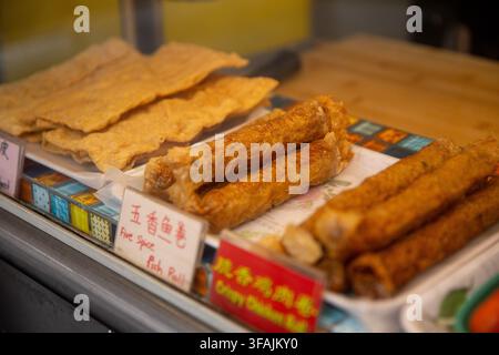 Trays of assorted fried tofu skin rolls, including Five Spice Fish Roll and Crispy Chicken Roll, displayed with handwritten labels at a Chinese street Stock Photo