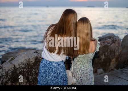 Sisters Looking at the Sea at Sunset. Two sisters standing together on a rocky shore and looking across the calm sea at sunset. Stock Photo