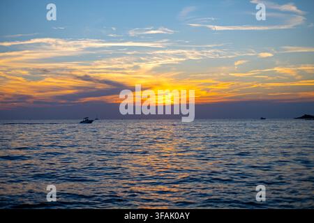 Golden Sunset Over Calm Sea with Boats. Orange sunset over calm sea with layered clouds, gentle waves, reflections and small boats silhouetted near Stock Photo
