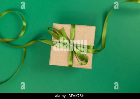 Top view of gift box wrapped in kraft paper with green satin ribbon bow on green background, showing loose ribbon curls Stock Photo