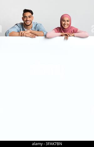 Nice Offer. Cheerful Muslim Spouses Leaning At Blank White Advertisement Board Stock Photo