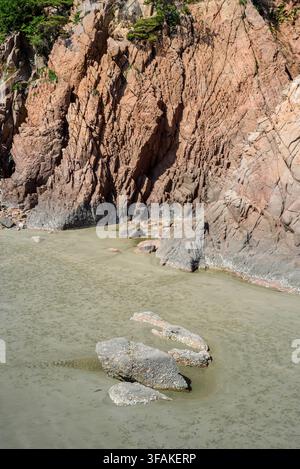 Coastal landscape dominated by a massive, rugged cliff face that rises steeply from a murky estuary. The composition features detailed Stock Photo