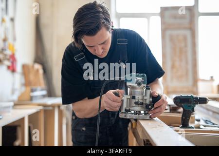 Craftsman using handheld wood router to remove layers from wooden board surface in repair studio. Stock Photo