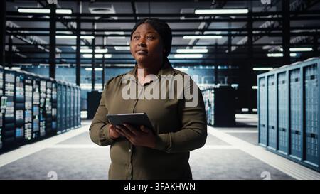 Admin in high tech data center using tablet to coordinate deployment of new server architecture. Server hub employee using device to do rollout of cybersecurity framework, protecting data, camera A Stock Photo