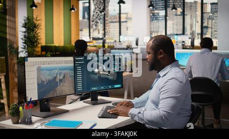 Technician using CAD software to optimize wind turbine blade aerodynamics, reducing carbon footprint. African american man in office testing advanced materials of eco friendly windmill, camera A Stock Photo