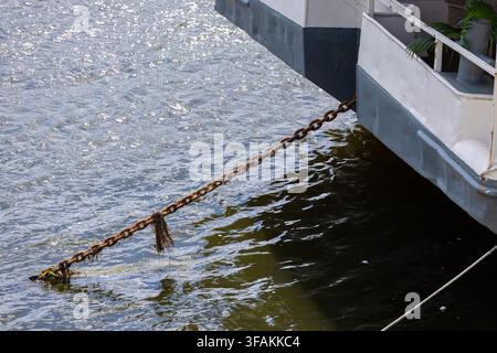 Ship stern with a rusted chain leading into rippled water. Seaweed rests on the metal links. White and gray railing sits above the stern. Stock Photo