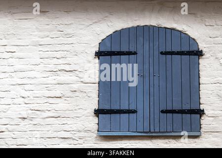 A dark wooden shutter with an arched top is mounted on a light brick wall. Vertical planks and metal hinges form a simple architectural detail. Stock Photo