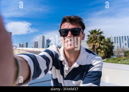 Young man in sunglasses taking a smiling selfie outdoors in a modern city with blue sky and palms Stock Photo