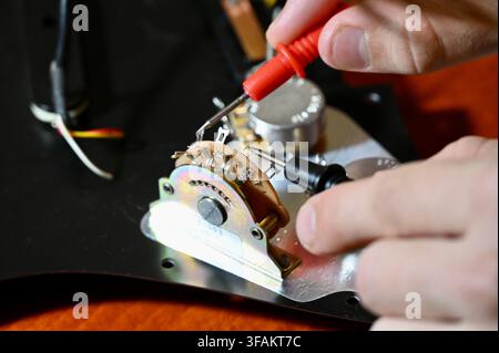 A commercial close-up photograph of a professional luthier carefully testing and diagnosing the electronic circuit of an electric guitar. The technician's hands are visible holding multimeter probes to check the wiring and components on a pickguard mounted on a wooden workbench. Stock Photo