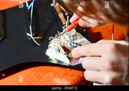 A commercial close-up photograph of a professional luthier carefully testing and diagnosing the electronic circuit of an electric guitar. The technician's hands are visible holding multimeter probes to check the wiring and components on a pickguard mounted on a wooden workbench. Stock Photo
