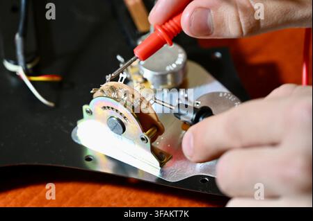 A commercial close-up photograph of a professional luthier carefully testing and diagnosing the electronic circuit of an electric guitar. The technician's hands are visible holding multimeter probes to check the wiring and components on a pickguard mounted on a wooden workbench. Stock Photo