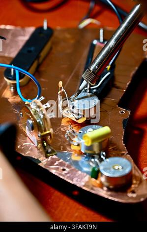 A commercial close-up photograph of a luthier soldering wiring and components on the copper-shielded control cavity of an electric guitar pickguard. A soldering iron with rising wisps of smoke is actively working on a potentiometer terminal, highlighting precise craftsmanship and repair. Stock Photo