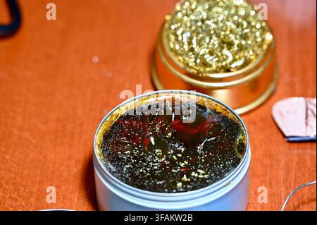 A commercial close-up photograph of an open round metal tin filled with dark, solid rosin flux used for electronics soldering and electrical work. The hard, amber-colored resin surface glistens under warm light, resting on a textured wooden workbench alongside other soldering tools and components. Stock Photo