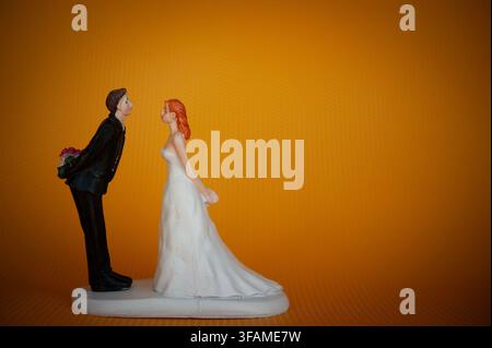 Wedding cake topper of a bride and groom facing each other, with the groom hiding flowers behind his back on a bright orange background Stock Photo
