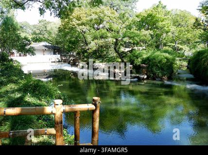 A bamboo railing frames a wide garden pond with a white traditional pavilion reflected in the still water, surrounded by rockery and lush trees. Stock Photo