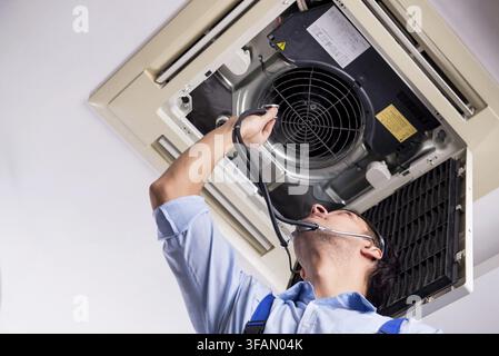 The young mechanic repairs the ceiling air conditioner Stock Photo