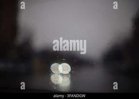 Raindrops on a car windshield creating a blurred background with soft glowing bokeh circles from street lamps during a rainy evening drive. Stock Photo