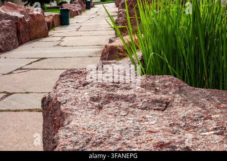 A paved walkway curves through a landscape featuring large granite boulders and vibrant green reeds Stock Photo