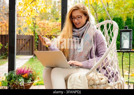 A mid adult woman works remotely on a laptop while drinking tea in a hanging swing chair on an autumn patio. Stock Photo